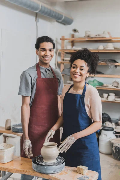 Smiling young african american couple shaping wet clay pot on wheel in pottery — Stock Photo