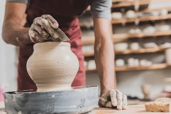 Partial view of young african american man in apron shaping wet clay pot on wheel with scraper in pottery — Stock Photo