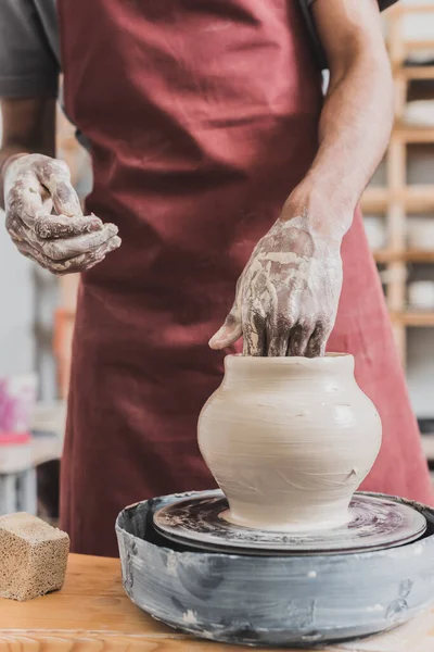 Partial view of young african american man in apron shaping wet clay pot on wheel with hands in pottery — Stock Photo