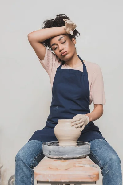 Cansada joven afroamericana en delantal sentada en el banco y moldeando la olla de arcilla húmeda con la mano cerca de la cabeza en cerámica - foto de stock