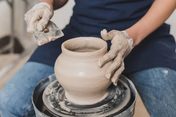 Partial view of young african american woman in apron sitting on bench and shaping wet clay pot with scraper in pottery — Stock Photo