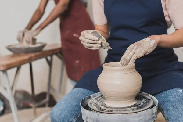 Partial view of young african american woman shaping wet clay pot on wheel with scraper in pottery — Stock Photo