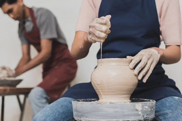 Vista parziale di giovane donna africana americana spremitura spugna mentre modellare pentola di argilla bagnata su ruota vicino uomo sfocato in ceramica — Foto stock