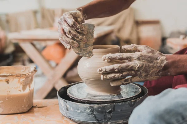 Крупным планом молодой африканский мужчина в фартуке лепит горшок на колесе с ломом в руке в керамике — стоковое фото