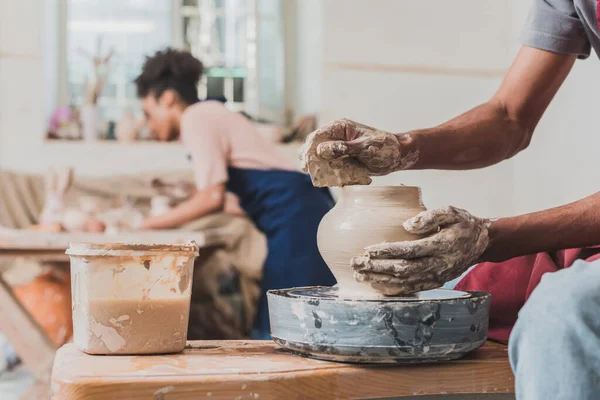 Young african american man sculpting pot on wheel with scraper in hand near blurred woman in apron in pottery — Stock Photo