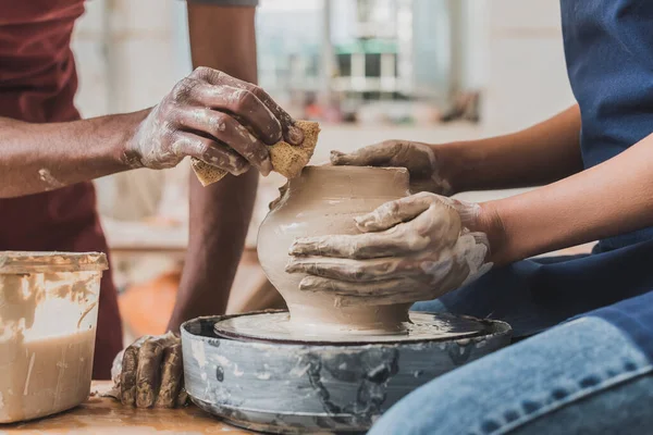 Partial view of young african american couple in aprons sculpting pot on wheel with wet sponge in pottery — Stock Photo