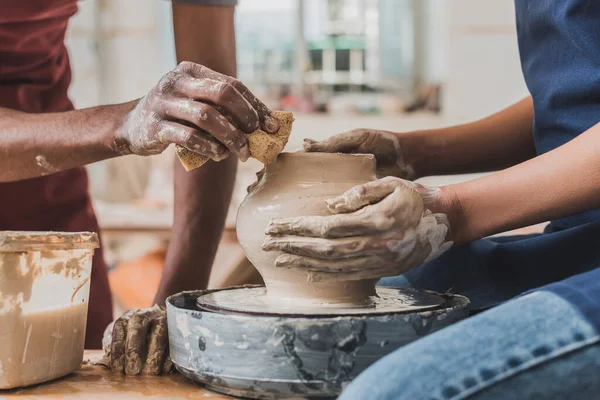 Vista parziale di giovane coppia afroamericana in grembiuli scultura pentola su ruota con spugna bagnata in ceramica — Foto stock