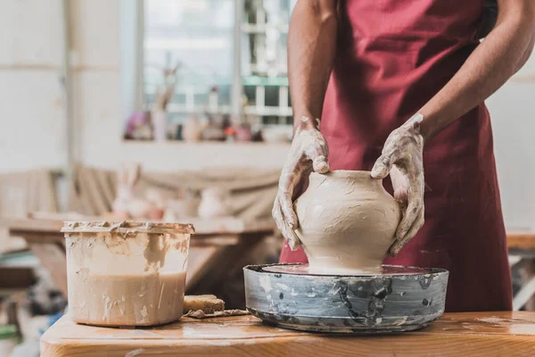 Partial view of young african american man sculpting pot on wheel near plastic box with water in pottery — Stock Photo