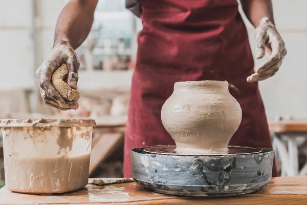 Vista parziale del giovane afroamericano in grembiule scolpire pentola di argilla su ruota e spremitura spugna bagnata in ceramica — Foto stock