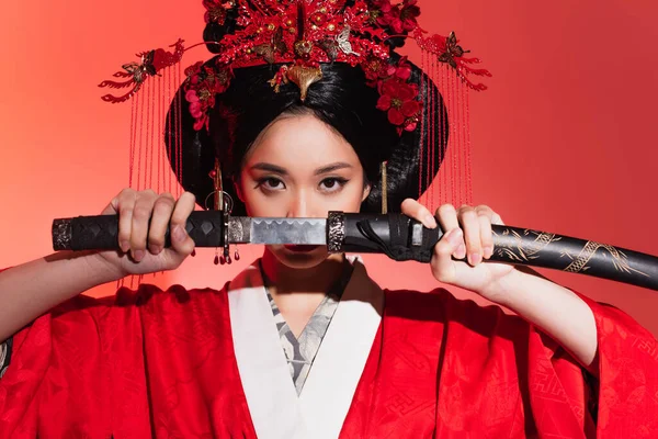 Joven mujer asiática cubriendo la cara con espada sobre fondo rojo - foto de stock