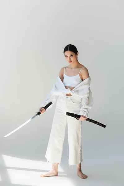 Barefoot japanese woman in white clothes holding sword on grey background — Stock Photo