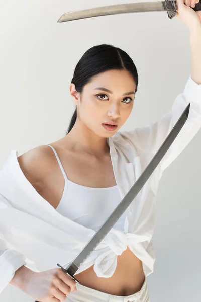 Jovem mulher japonesa de camisa branca segurando espadas isoladas em cinza — Fotografia de Stock