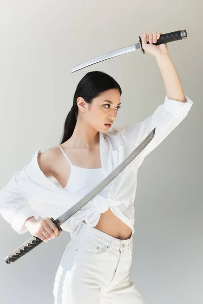 Femme asiatique avec des épées regardant loin dans la lumière du soleil isolé sur gris — Photo de stock