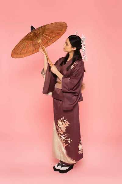 Side view of asian woman in traditional clothes holding umbrella on pink background — Stock Photo