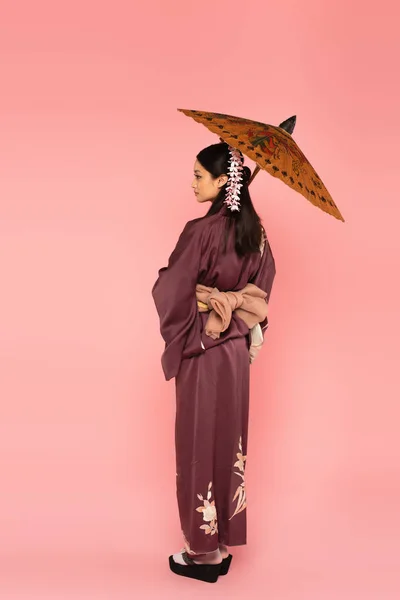 Вид збоку азіатки в східному одязі, що тримає парасольку на рожевому фоні — стокове фото