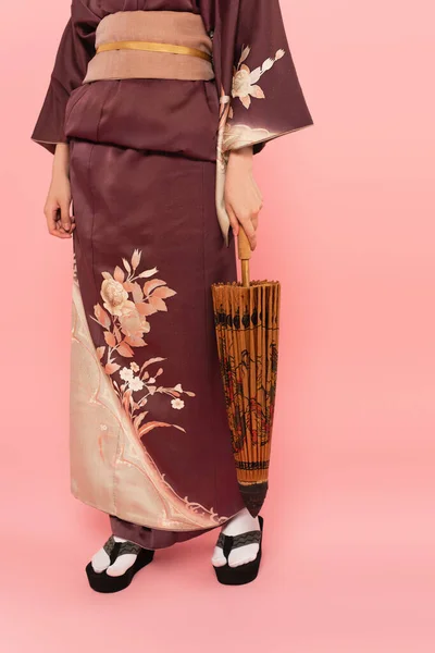 Vista ritagliata della donna in vestiti giapponesi e calzature che tengono l'ombrello su sfondo rosa — Foto stock