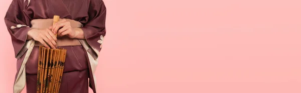 Обрізаний вид молодої жінки в кімоно тримає парасольку ізольовано на рожевому, банер — стокове фото