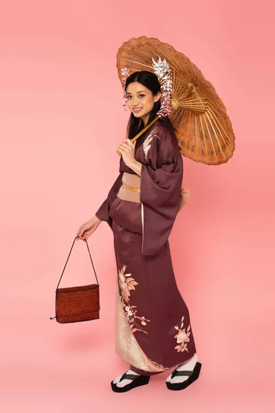 Smiling asian woman holding umbrella and handbag on pink background — Stock Photo
