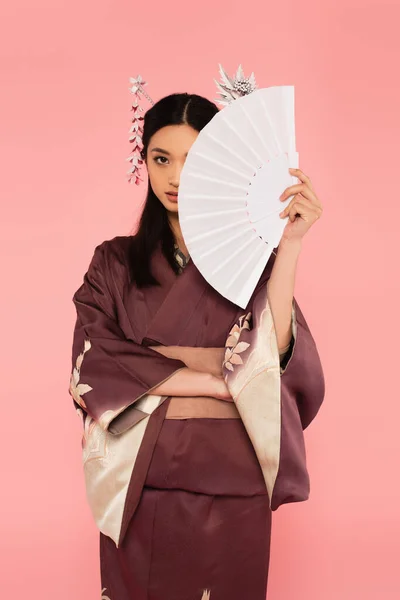 Mulher asiática em quimono segurando ventilador perto do rosto isolado em rosa — Fotografia de Stock