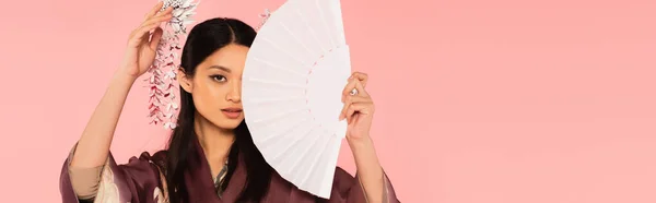 Femme asiatique avec coiffure traditionnelle tenant ventilateur près du visage isolé sur rose, bannière — Photo de stock