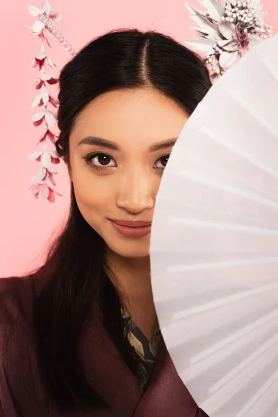 Junge asiatische Frau schaut in die Kamera, während sie Ventilator isoliert auf rosa hält — Stockfoto