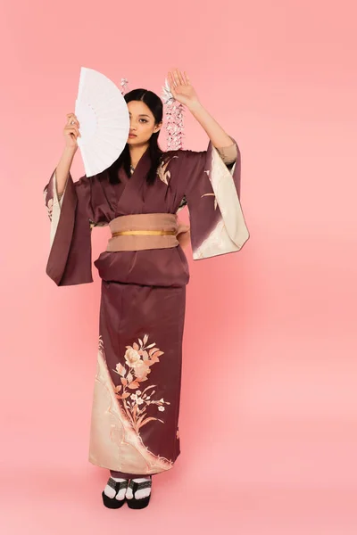 Lunghezza completa della donna giapponese in kimono che tiene il ventilatore vicino al viso su sfondo rosa — Foto stock