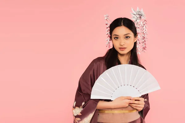 Mulher asiática bonita segurando ventilador branco e olhando para câmera isolada em rosa — Fotografia de Stock