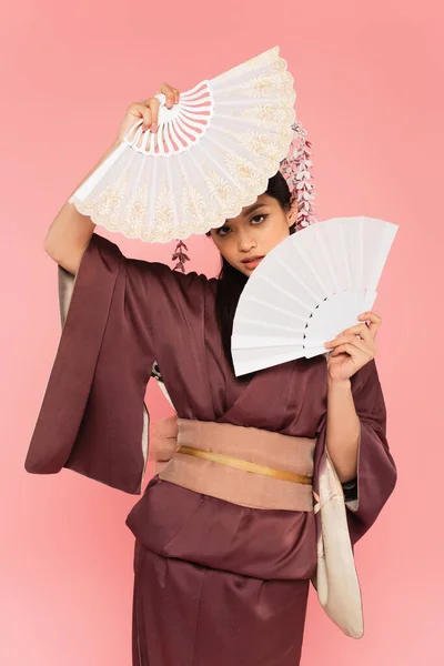Giovane donna asiatica che tiene i fan vicino al viso isolato sul rosa — Foto stock