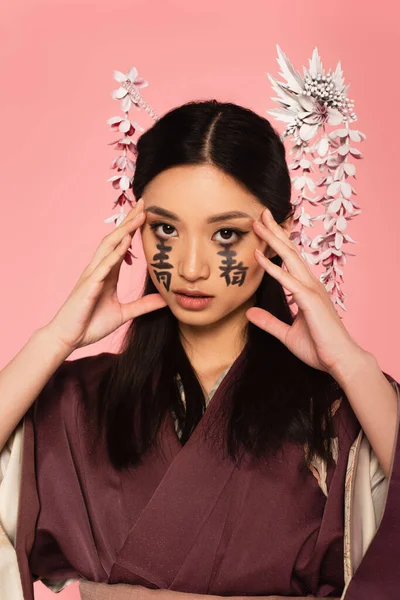 Asian woman with hieroglyphs on face looking at camera isolated on pink — Stock Photo