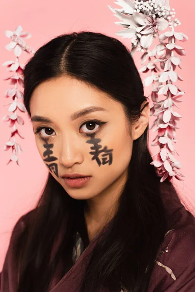 Jeune femme asiatique avec hiéroglyphes sur le visage regardant la caméra isolée sur rose — Photo de stock