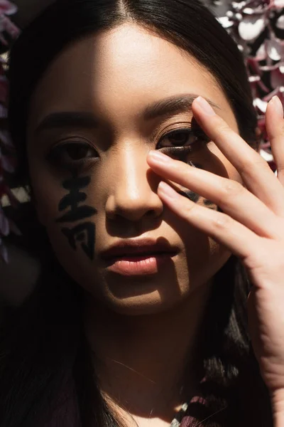 Femme asiatique avec hiéroglyphes sur le visage regardant la caméra dans la lumière isolée sur noir — Photo de stock