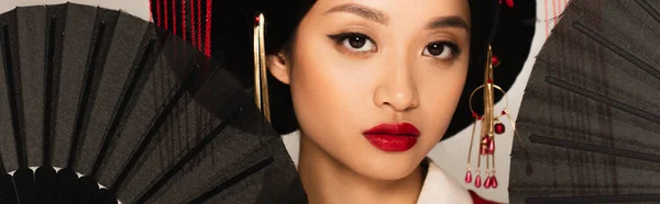 Mujer asiática con labios rojos mirando a la cámara cerca de ventiladores negros aislados en gris, pancarta - foto de stock