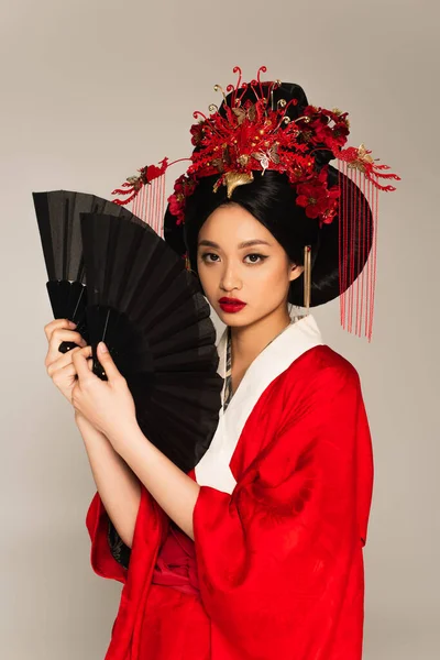 Bonita mujer asiática en kimono rojo sosteniendo ventiladores y mirando a la cámara aislada en gris - foto de stock