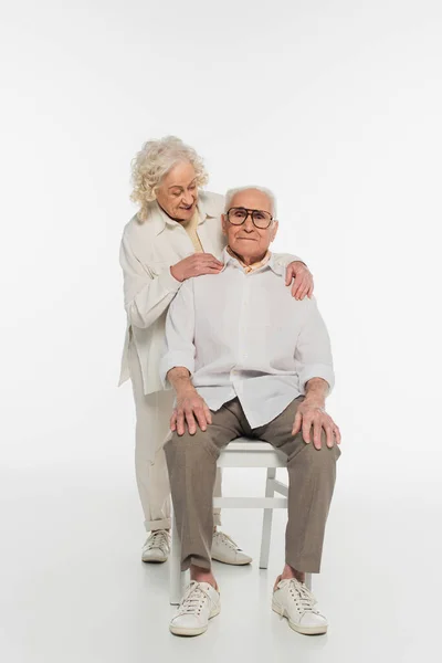 Elderly woman gently hugging husband sitting on chair on white — Stock Photo