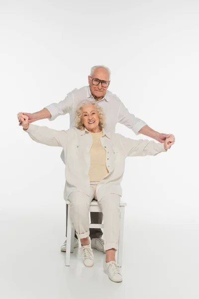 Happy elderly man holding outstretched hands of wife sitting on chair on white — Stock Photo