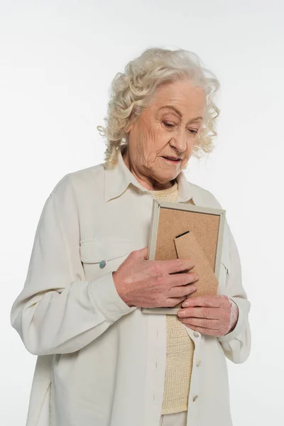 Triste mulher idosa em blusa e camisa segurando moldura foto perto do peito isolado no branco — Fotografia de Stock