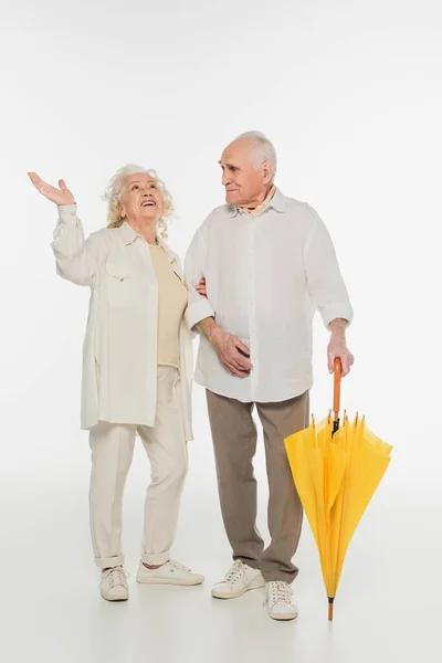 Amazed elderly woman with hand in air standing with husband holding umbrella on white — Stock Photo