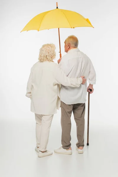 Visão traseira do casal de idosos em roupas casuais andando com guarda-chuva amarelo e vara no branco — Fotografia de Stock