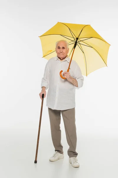 Elderly man in casual clothes standing with yellow umbrella and walking stick on white — Stock Photo