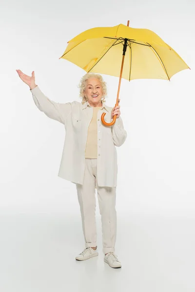 Anciana sonriente en ropa casual con gesto de triunfo de pie con paraguas amarillo sobre blanco — Stock Photo