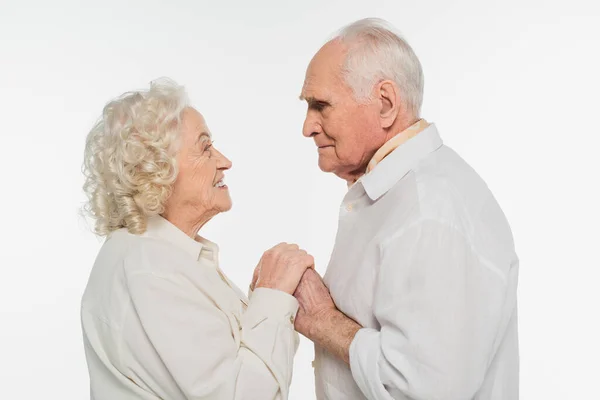 Feliz pareja de ancianos suavemente tomados de la mano y mirándose unos a otros aislados en blanco - foto de stock