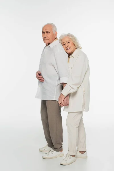 Happy elderly woman gently hugging back of husband with holding hands and looking at camera on white — Stock Photo