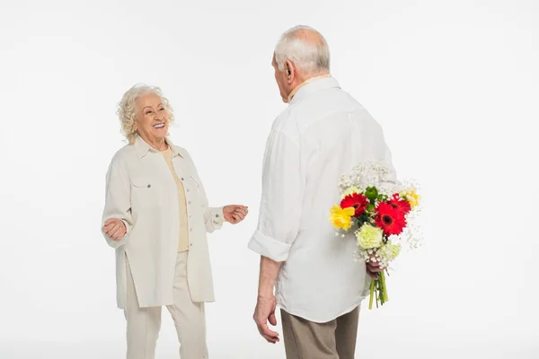 Homme âgé cachant bouquet de fleurs derrière le dos près de la femme avec les mains tendues sur blanc — Photo de stock