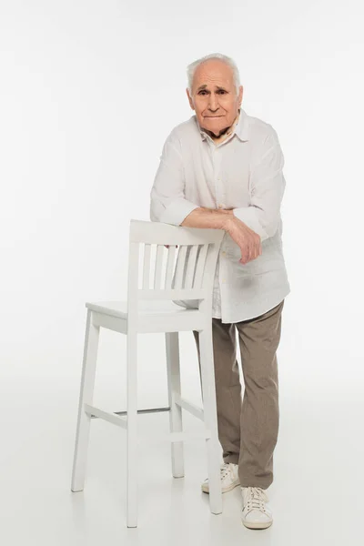 Upset elderly man in casual clothes standing near stool and looking at camera on white — Stock Photo