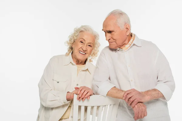 Feliz pareja de ancianos en ropa casual de pie cerca de taburete aislado en blanco - foto de stock