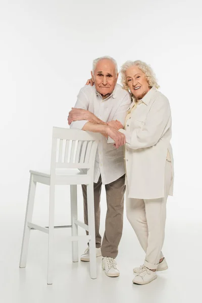 Счастливая пожилая пара в повседневной одежде, стоящая возле стула и обнимающаяся на белом — стоковое фото