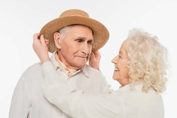 Sonriente anciana ajustando sombrero en la cabeza del marido aislado en blanco - foto de stock