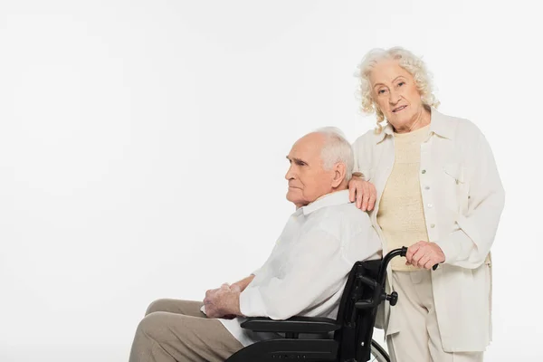 Mulher idosa de pé perto do marido em cadeira de rodas isolada em branco — Fotografia de Stock