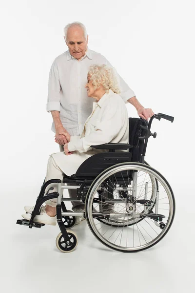 Sad elderly woman in wheelchair holding hand of husband on white — Stock Photo