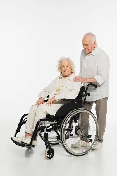 Elderly man holding hand on shoulder of wife in wheelchair isolated on white — Stock Photo